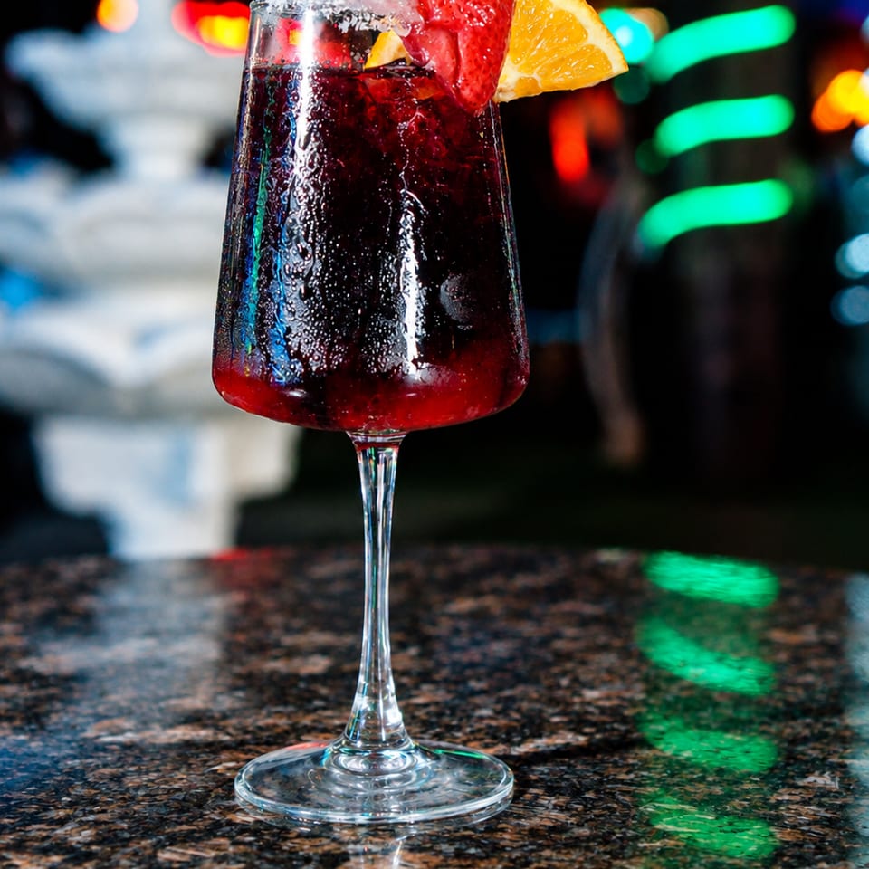 Dark red cocktail in a sugar-rimmed wine glass, garnished with strawberries and an orange wedge on a reflective bar.