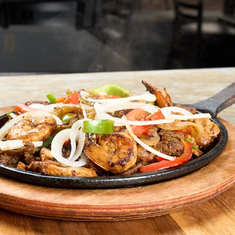 Sizzling skillet of mixed fajitas with shrimp, steak, chicken, peppers, and onions on a wooden board.