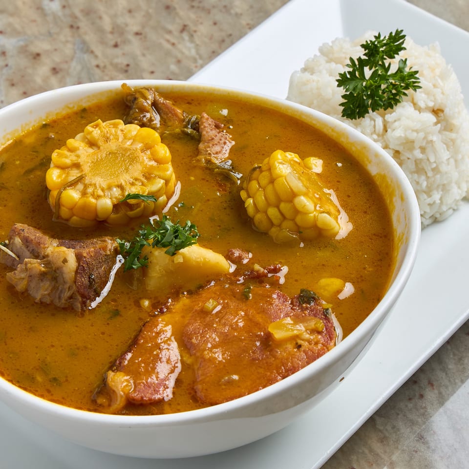 Bowl of richly seasoned stew with meat, corn, potatoes, and vegetables, served alongside white rice garnished with parsley.
