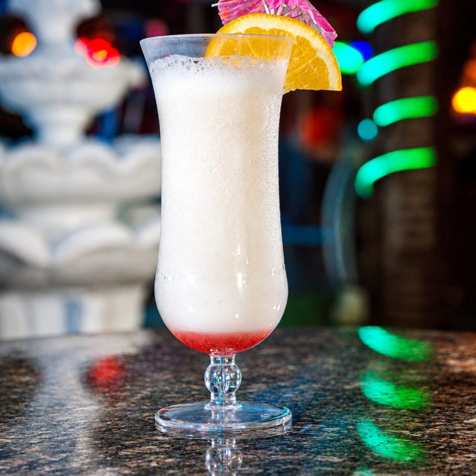 Creamy white cocktail with a red base, orange slice, and pink umbrella in a goblet on a reflective bar.