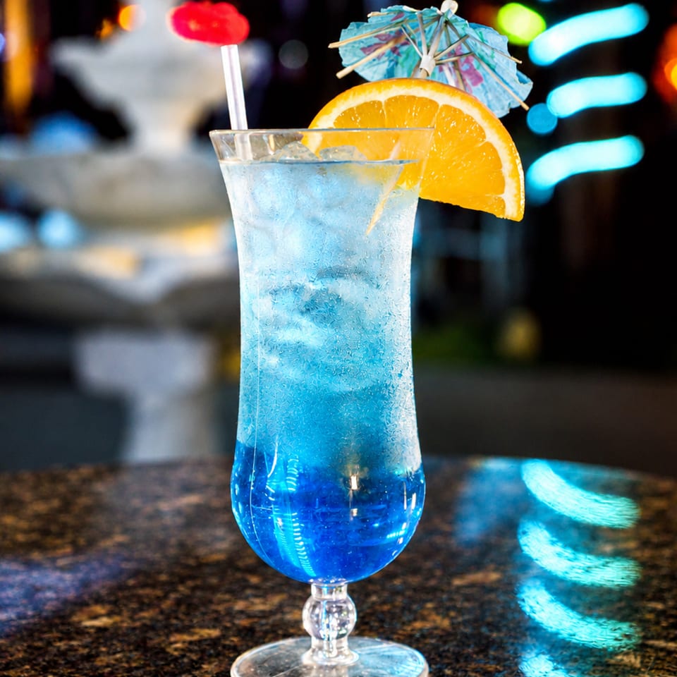 Bright blue cocktail in a tall glass garnished with an orange slice, blue umbrella, and red lollipop on a bar counter.